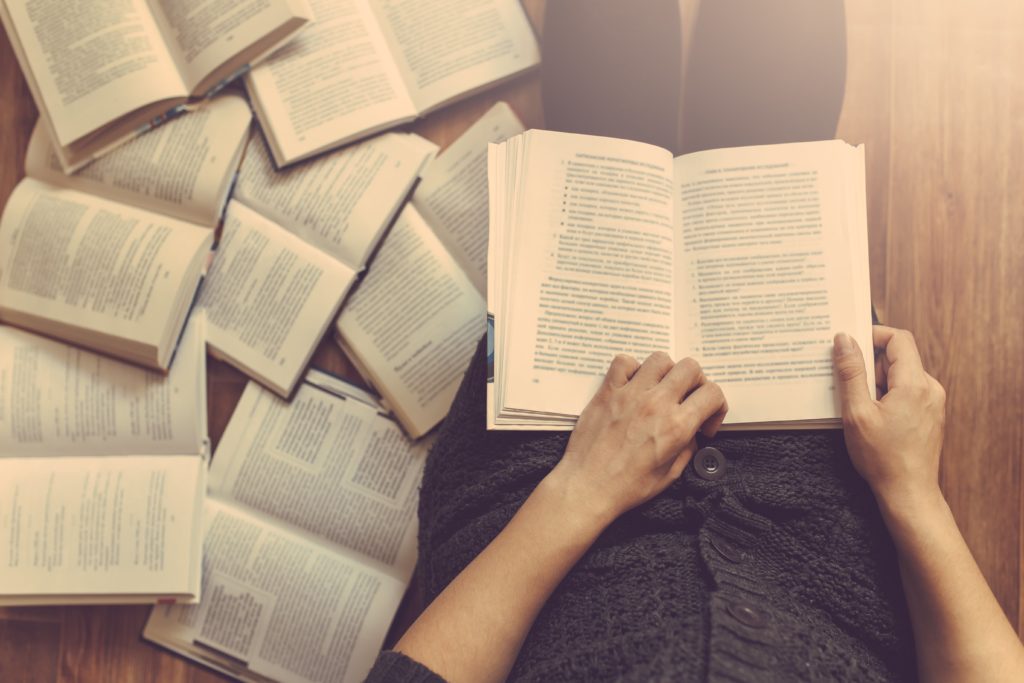 Woman sitting on a wooden floor reading a book with open books laying next to her.